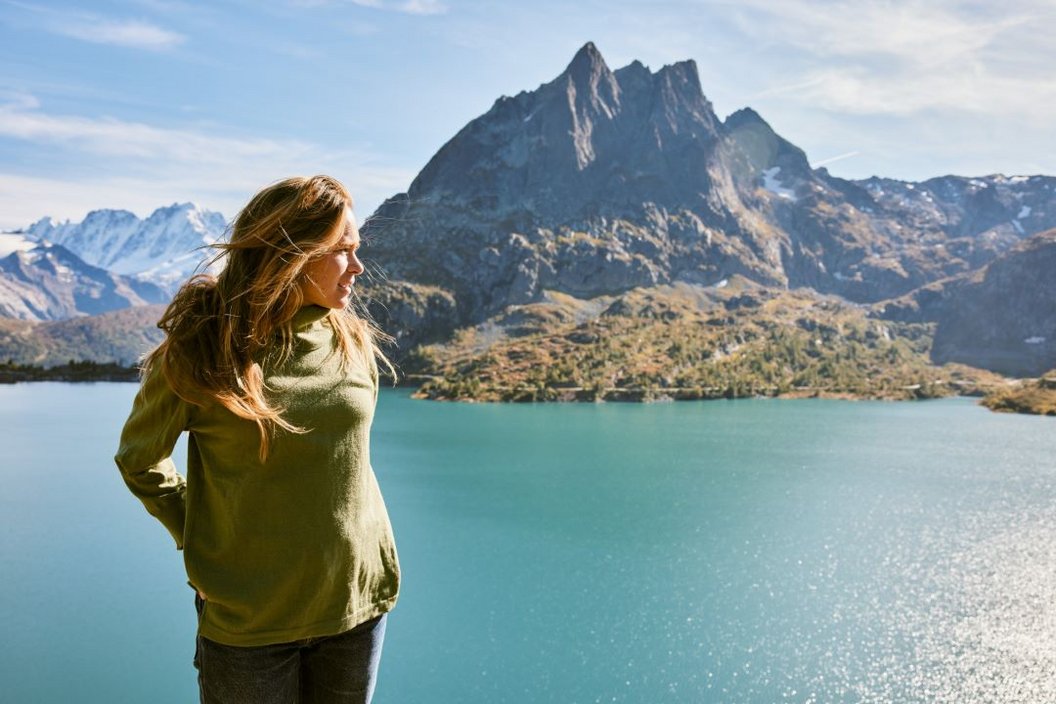 Woman is looking to the side and behind her is a beautiful blue lake with a mountain towering over it