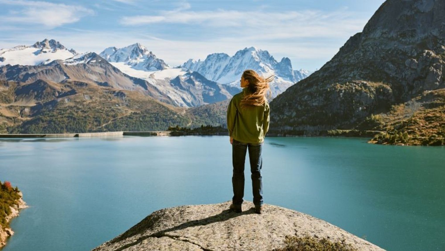 Woman on rock staring at mountains and lake A woman is standing on a rock with a big blue mountain lake beneth hair. She is looking to a vast mountain landscape
